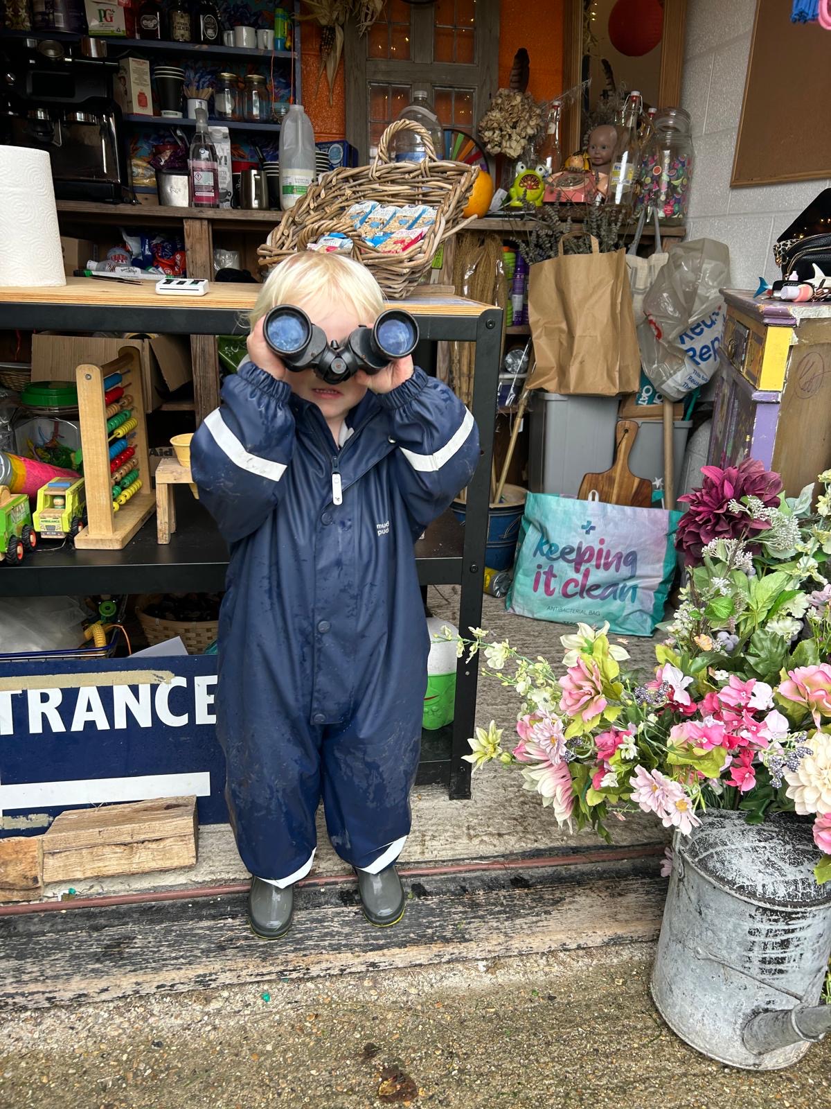 A child in a muddy navy puddlesuit looking through big binoculars